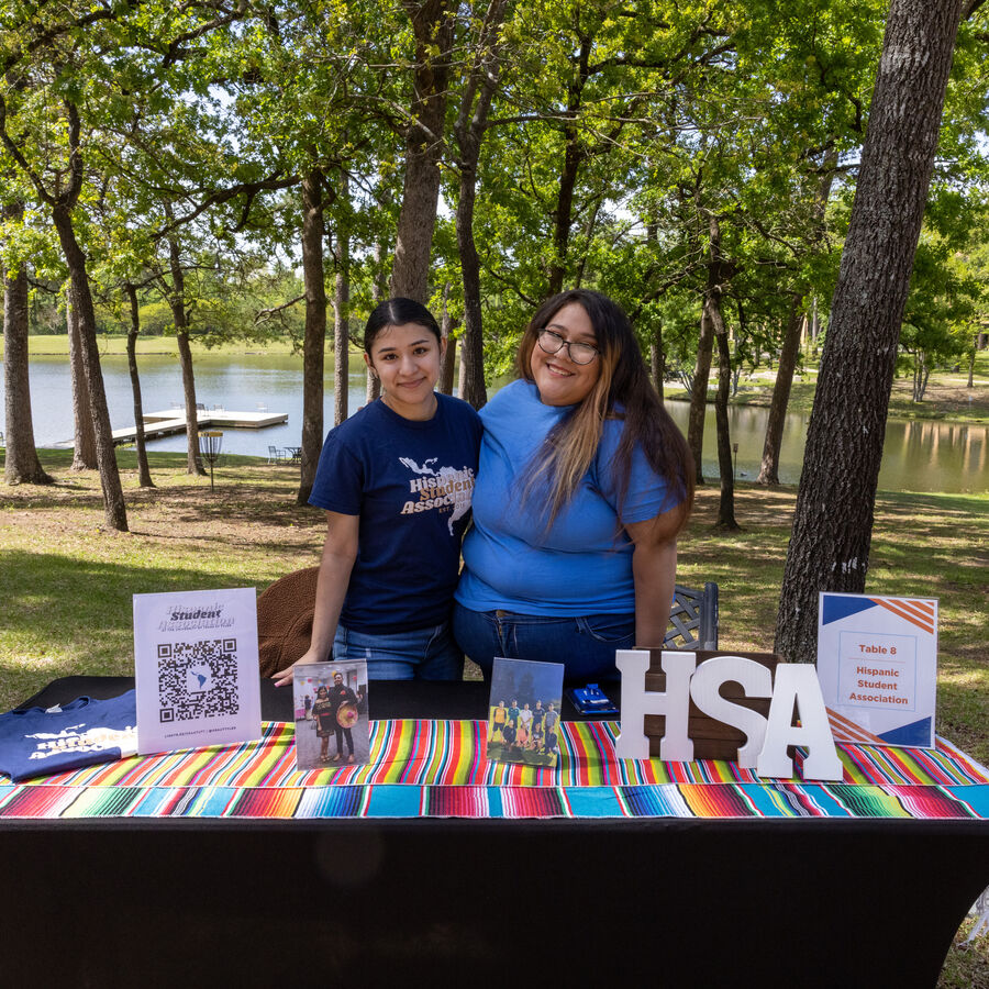 Two University of Texas at Tyler students stand in front of an outdoor table display for the Hispanic Student Association