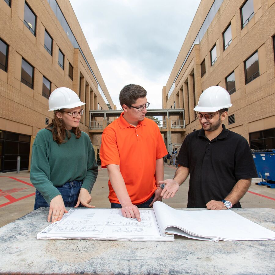 Three students, two male and one female, standing between two brick buildings looking at construction plans.