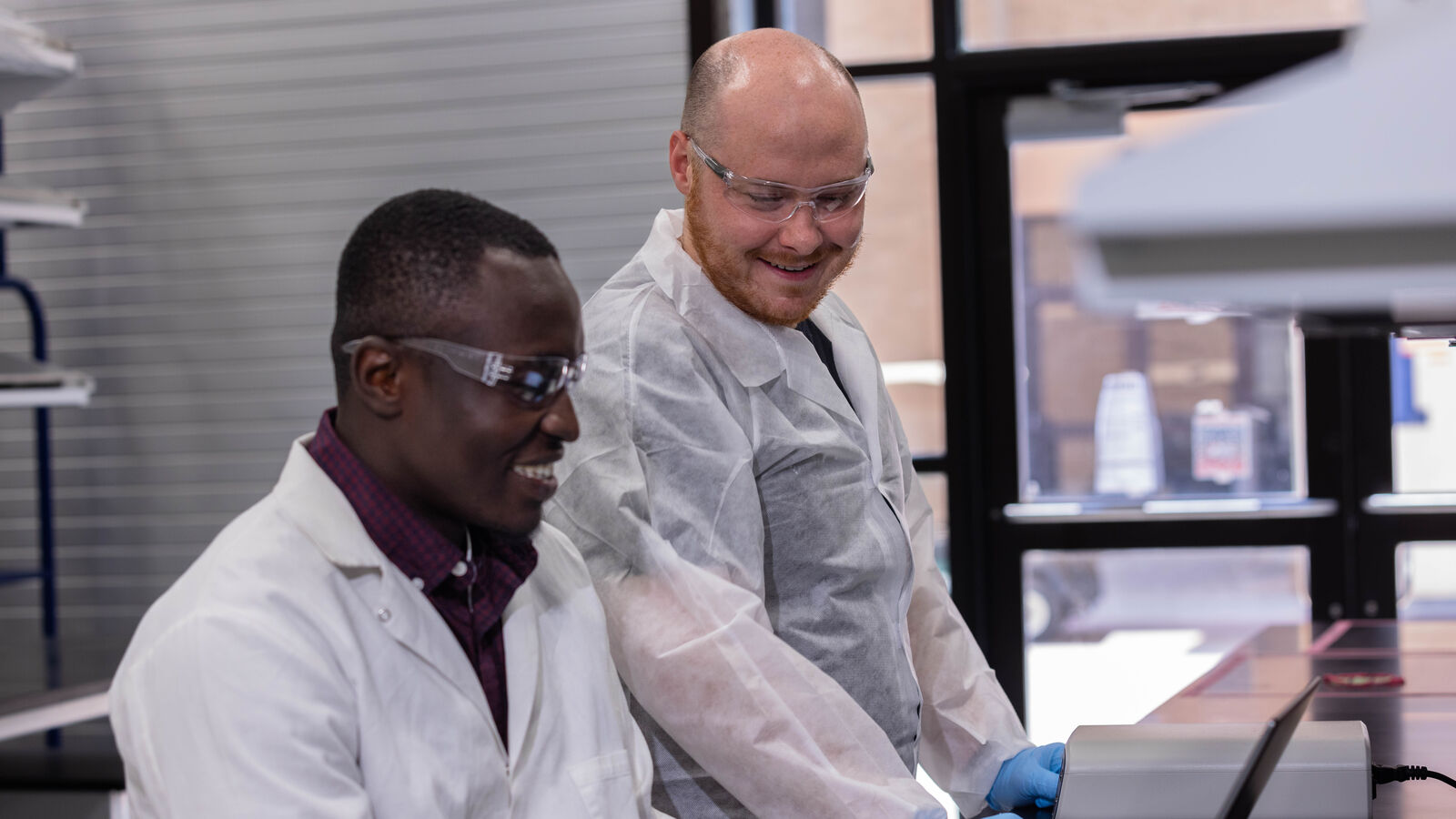 Two MS in Biotechnology students use a laptop while wearing lab coats and safety glasses in the chemical engineering lab at UT Tyler