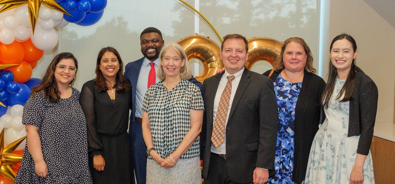 "Group photo of seven individuals dressed formally, standing in front of a colorful balloon arch featuring blue, orange, white, and gold balloons. Gold number balloons are visible in the background, and a window with trees outside adds natural light to the setting.