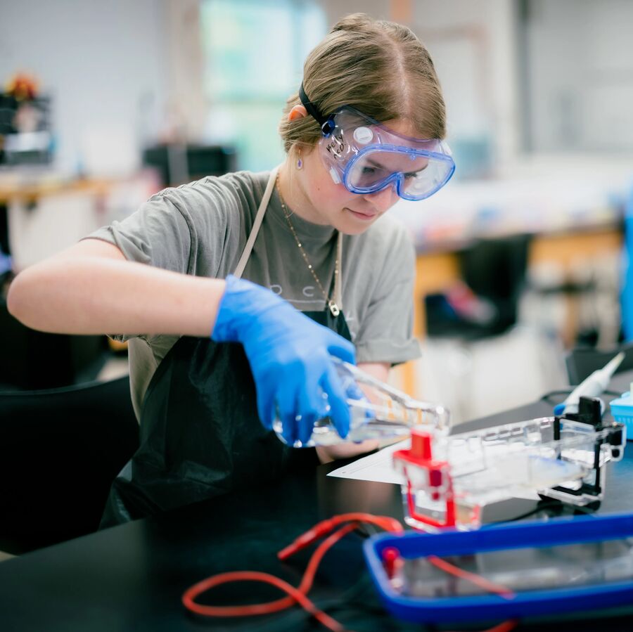 A young student wearing protective gear in a science classroom at the UT Tyler Palestine University Academy pours liquid from a beaker.