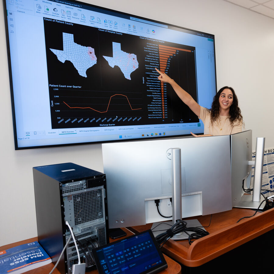A woman points at graphs on a large screen displaying healthcare data.
