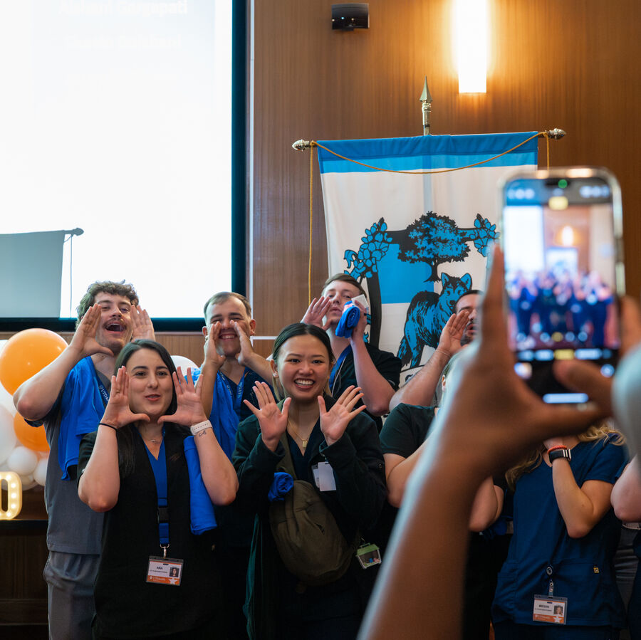 A group of people in scrubs poses playfully indoors with a banner and balloons in the background. A smartphone in the foreground is capturing the scene.