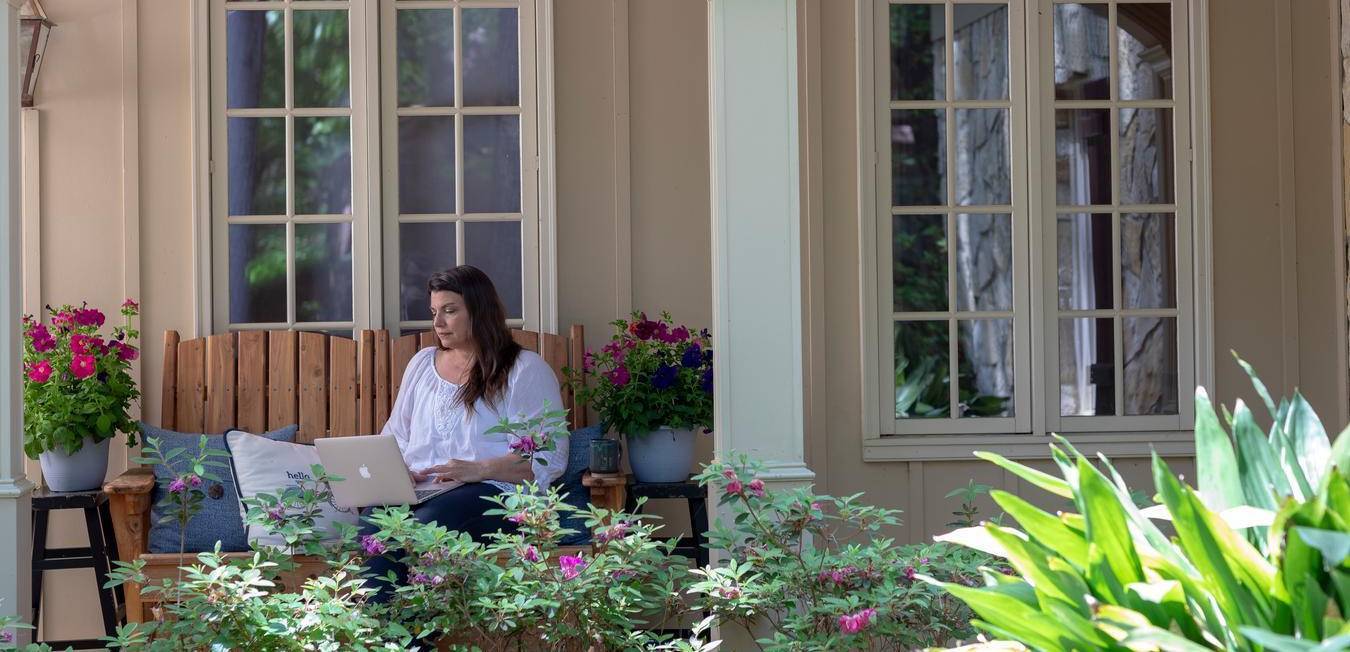 A lady sitting on a brown chair, surrounded by flowers working on a laptop