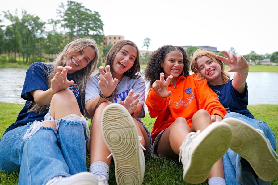 Four students sitting in the grass holding up the talons handsign